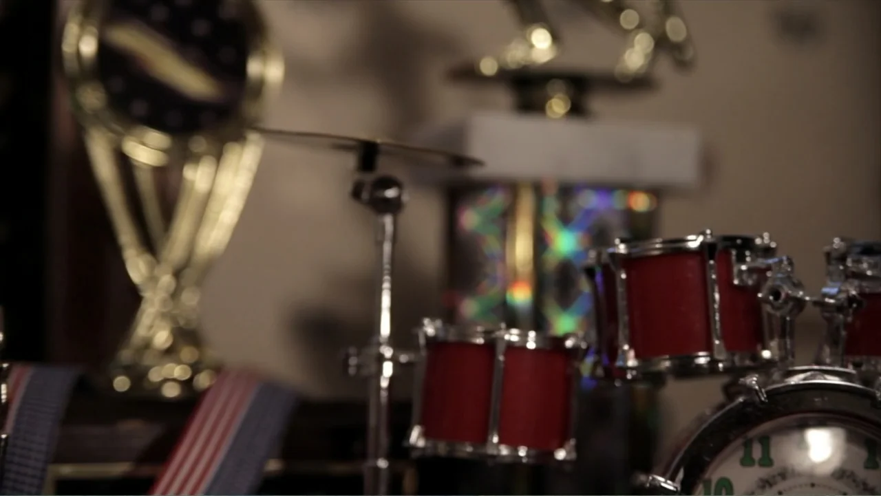 Life According to Sam (2013) movie still: closeup — A collection of personal items sits on a surface inside a dimly lit room. Miniature drums and…
