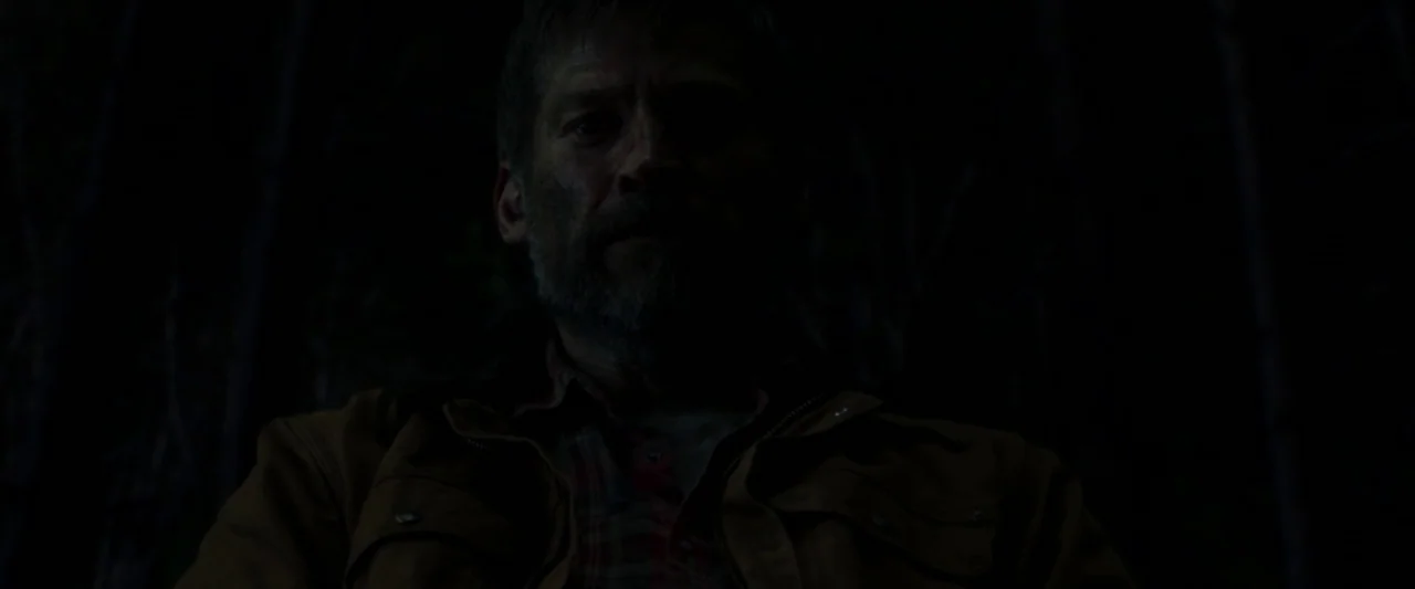 The Silencing (2020) movie still: medium — A man stands in a dark environment with tall, indistinct vertical shapes behind him. The man looks…