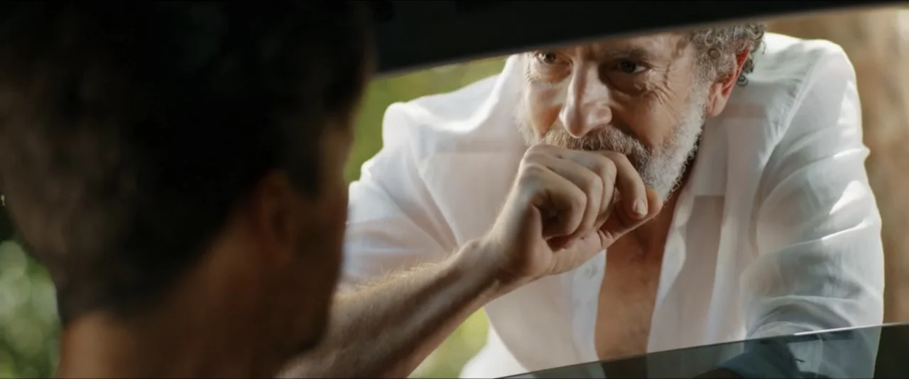 Gatlopp: Hell of a Game (2022) movie still: medium — An older man peers into the interior of a vehicle from outside. He rests his elbows on the…