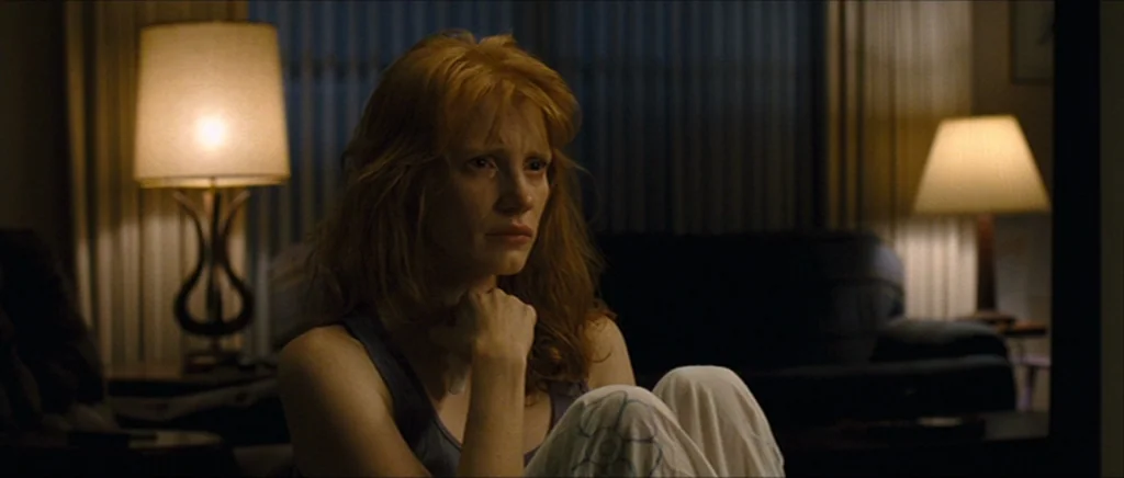 Take Shelter (2011) movie still: medium — A dimly lit room features a woman sitting on the floor by a sofa. She clutches her knees while facing…
