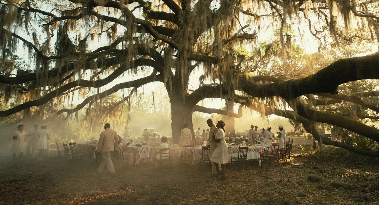 The Color Purple (2023) movie still: wide — A group of people in 1950s attire attend a celebration under a massive oak tree. Long banquet tables…