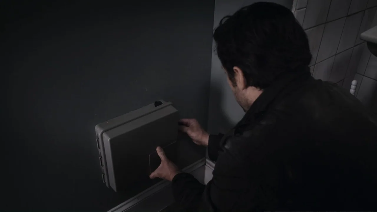 Flypaper (1999) movie still: medium — A dimly lit utility room features a grey plastic junction box mounted on dark walls. A man in a dark jacket…