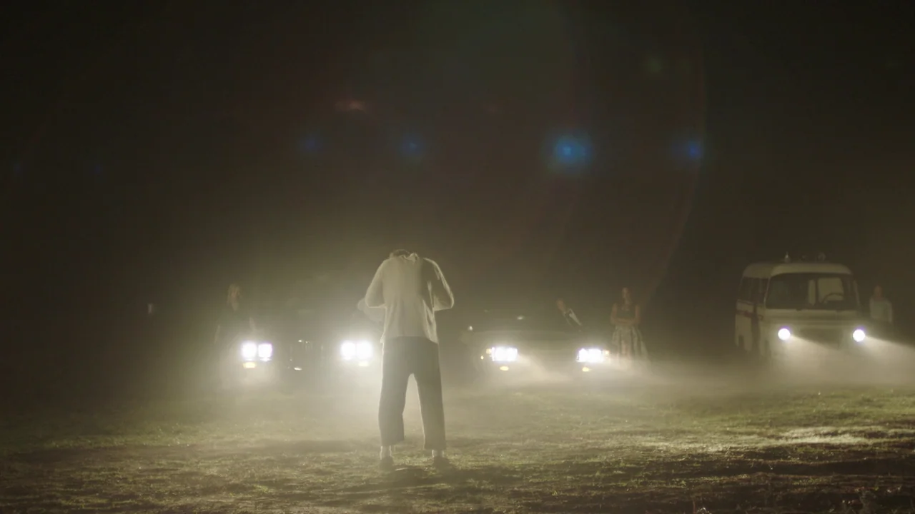 Infinity Pool (2023) movie still: wide — A dark, foggy open field is illuminated by the bright headlights of three parked vehicles. A person stands…