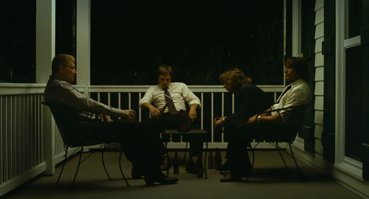 The Iron Claw (2023) movie still: wide — A dark, wood-railed porch at night with four men sitting in chairs. The men are grouped around a small…