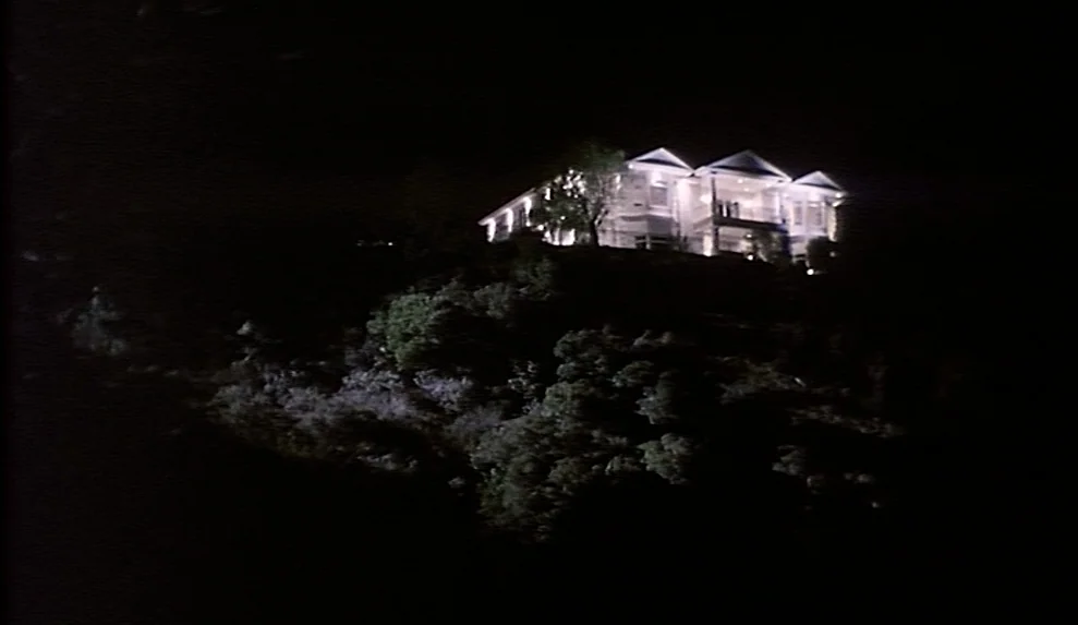 Safe movie still: establishing — A multi-level white house is perched on a dark, tree-covered hillside at night. Bright lights shine from the…