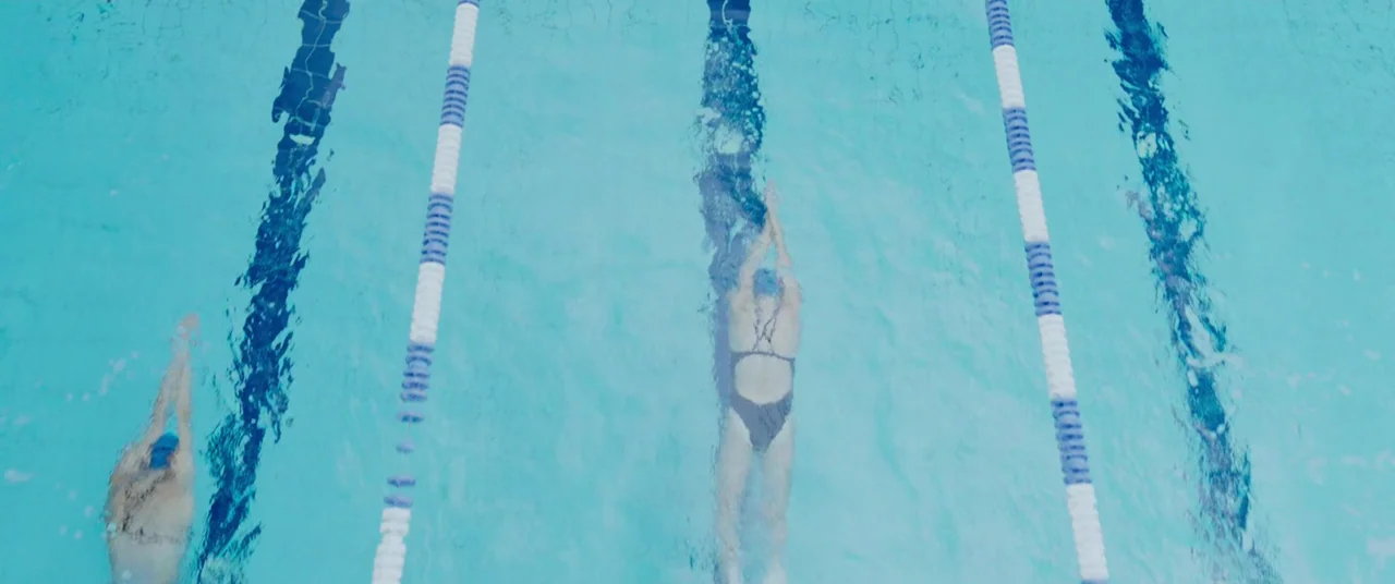 Crawl (2019) movie still: wide — An outdoor swimming pool with bright blue water and parallel lane dividing lines. Two swimmers glide through the…
