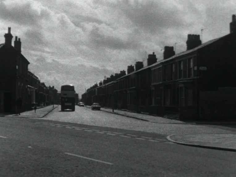Children (1976) movie still: establishing — A long row of Victorian-style terrace houses lines a paved street under cloudy skies. A double-decker…