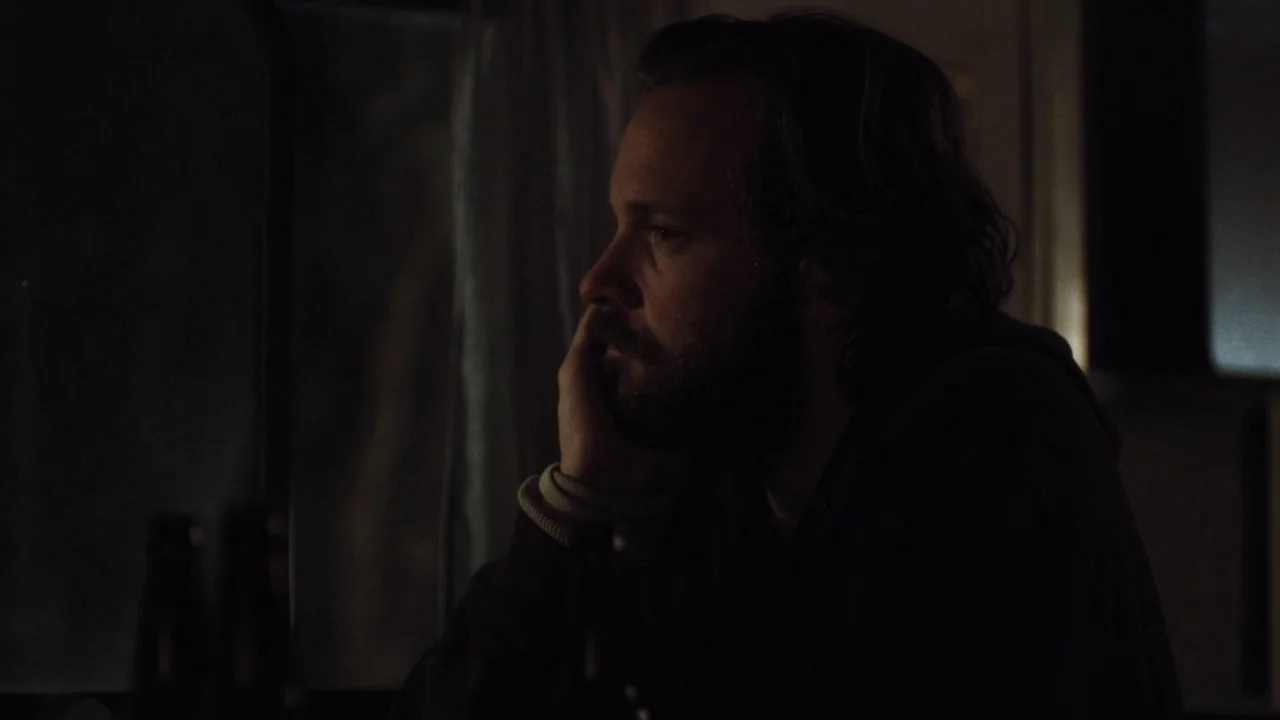 Night Moves (2013) movie still: closeup — A dark indoor space with a window draped by thin curtains. A man sits in profile, resting his hand against…