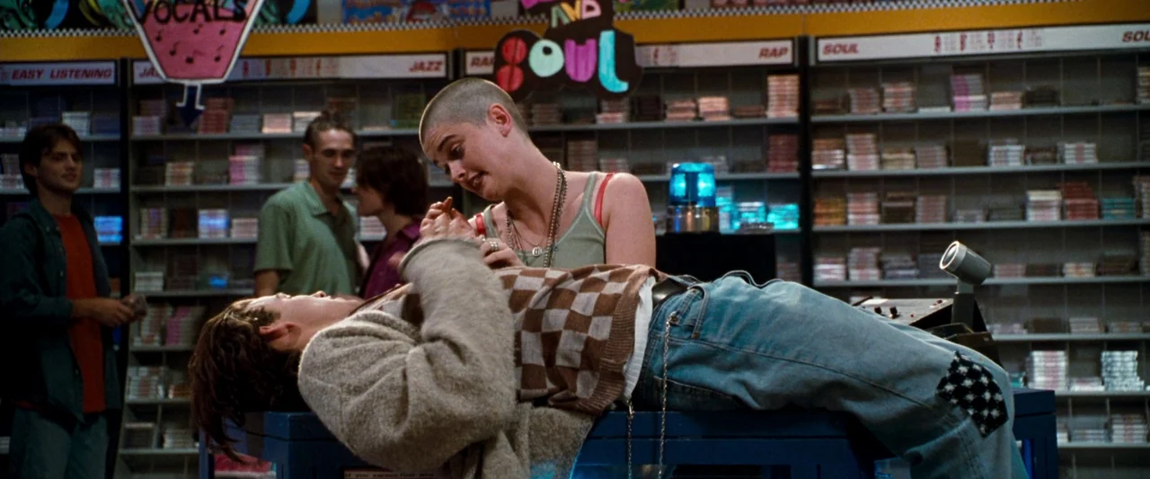 Empire Records (1995) movie still: medium — A music store interior is lined with shelves containing numerous rows of cassette tapes. A young woman…