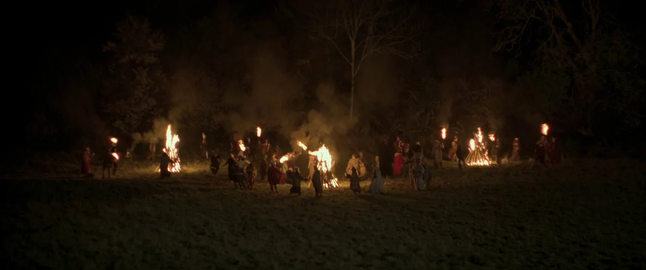 Lord of Misrule (2023) movie still: wide — A dark night landscape features a large group of people gathered in an open field. Several large bonfires…