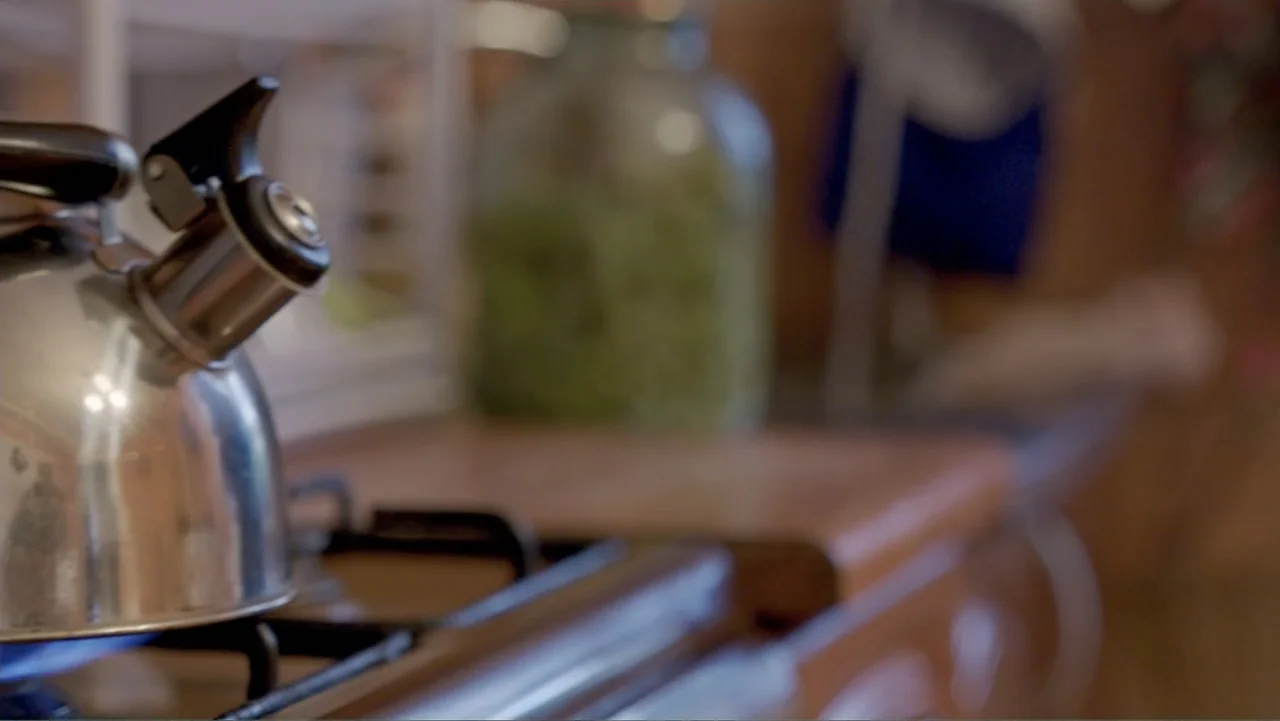 Finding the Way Home (1991) movie still: closeup — A close view of a stainless steel tea kettle on a stovetop. A large glass jar containing green…