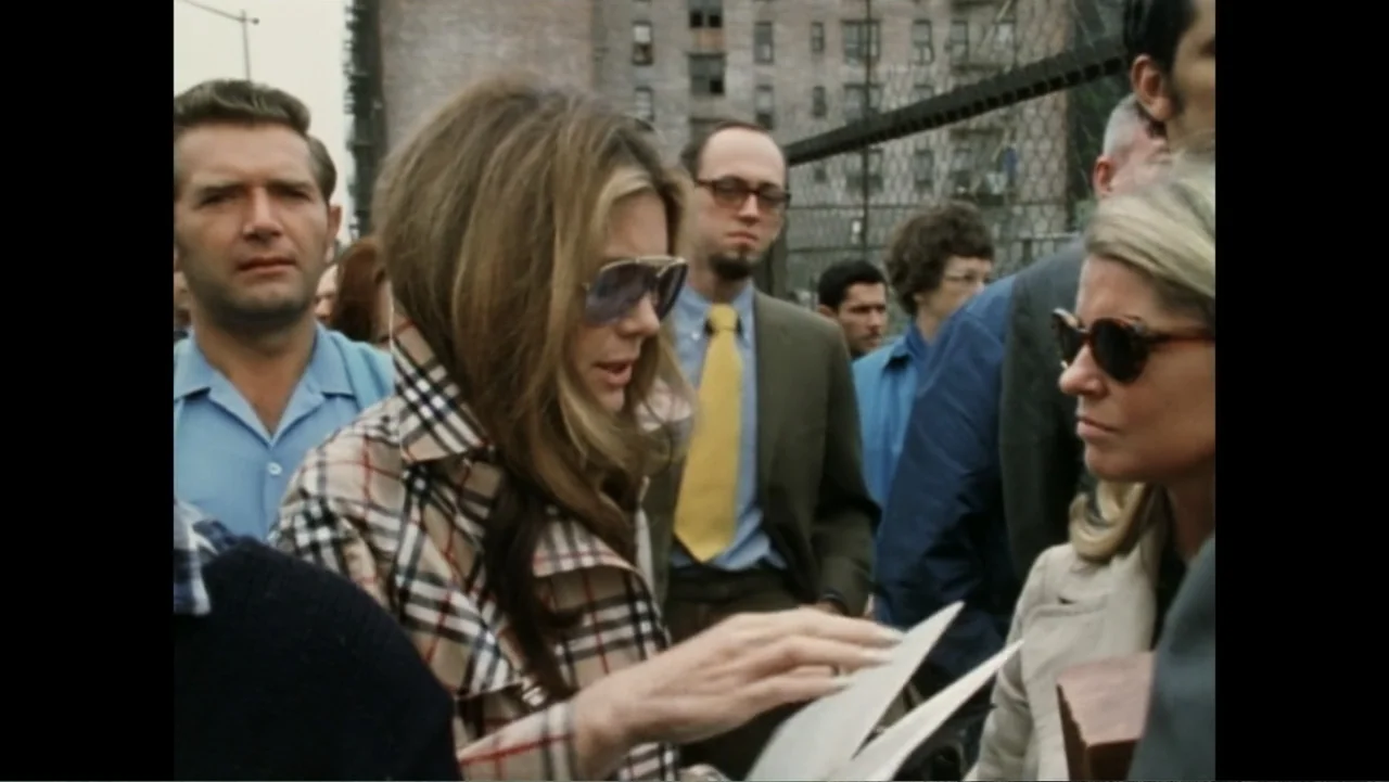 Dear Ms.: A Revolution in Print (2025) movie still: medium — A crowded urban sidewalk outside a chain-link fence with tall brick buildings behind. A…