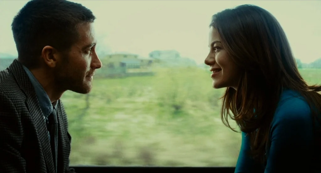 Source Code (2011) movie still: medium — A man and a woman face each other inside a train car with a window. Blurred green countryside passes by…