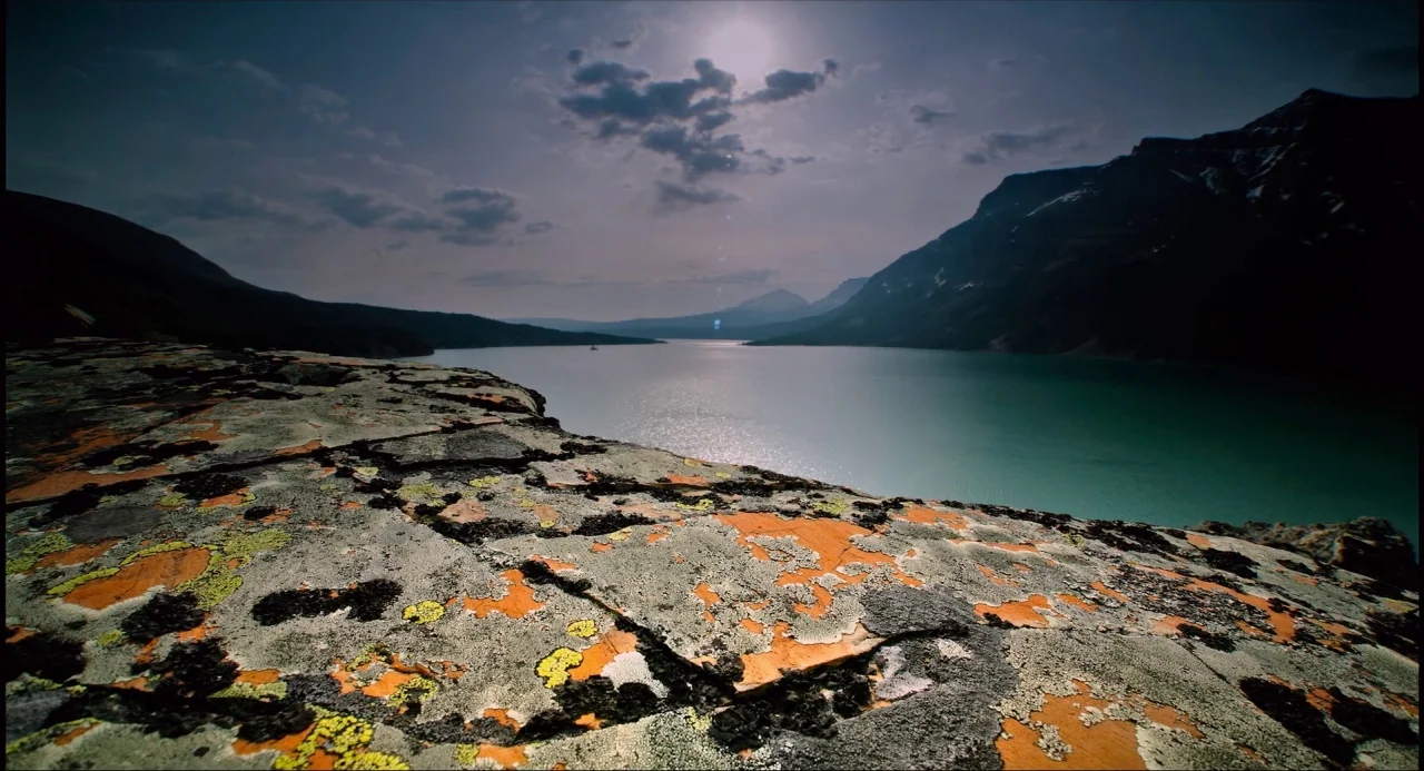 Voyage of Time (2016) movie still: establishing — A lichen-covered rocky foreground overlooks a calm, wide lake surrounded by distant dark…