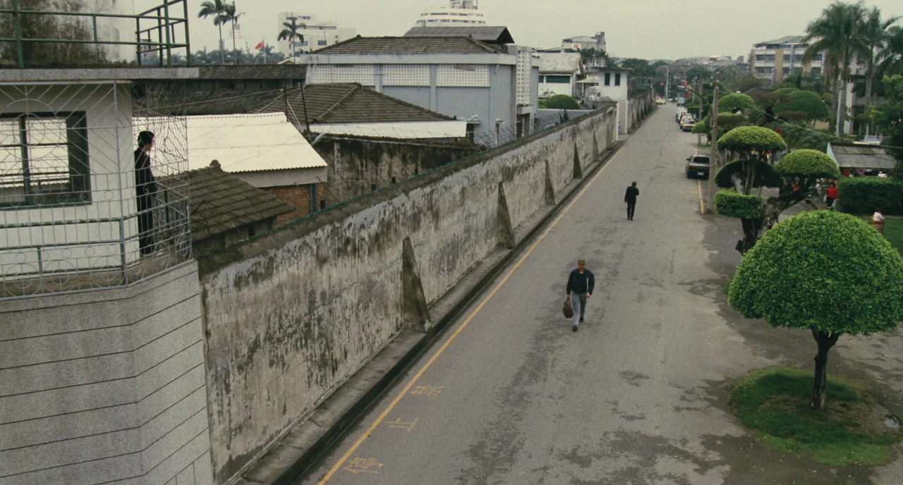 A Better Tomorrow (1986) movie still: wide — A long, weathered concrete wall stretches along a paved road lined with trimmed trees. A man walks down…