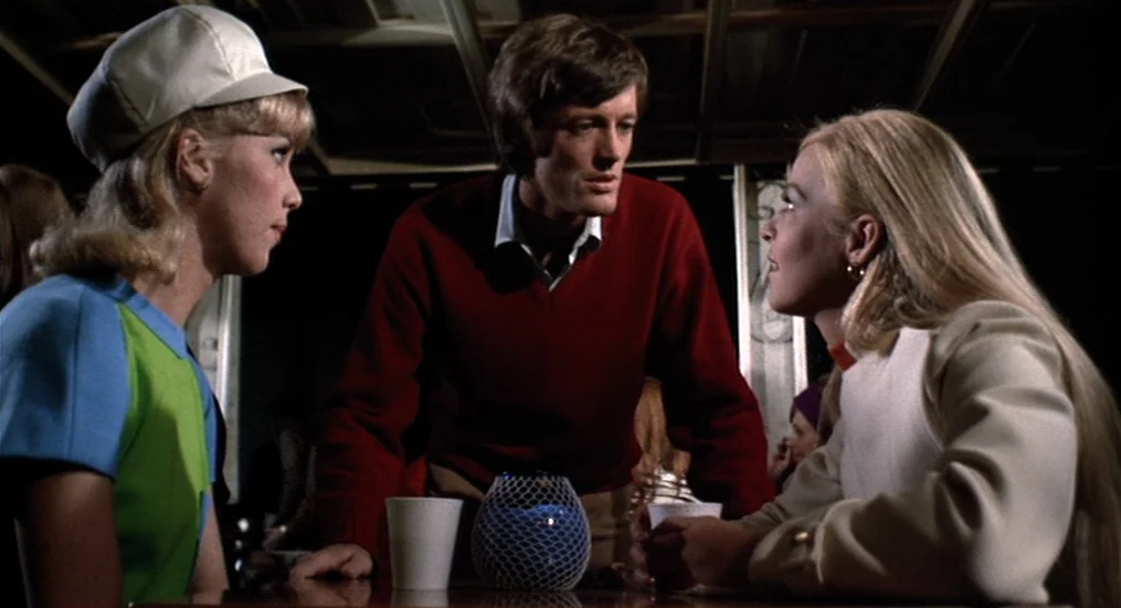 The Trip (1967) movie still: medium — A dark indoor space features a booth with three young adults engaged in conversation. The man stands in the…