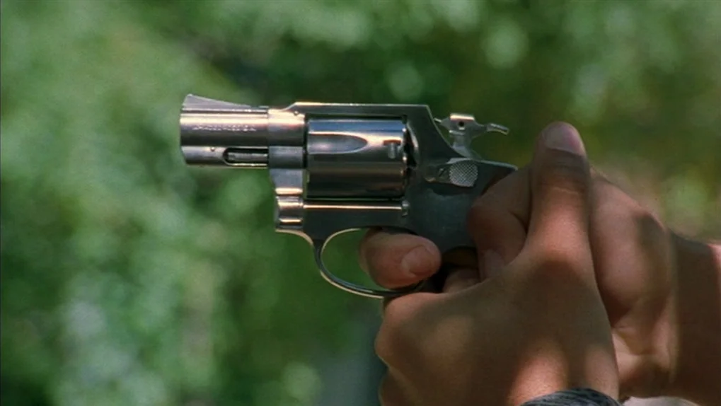 Mean Creek (2004) movie still: closeup — A silver snub-nosed revolver is held by two hands against a blurred green background. The metallic frame…