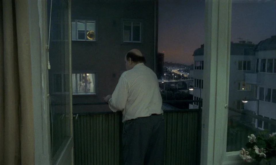 You, The Living (2007) movie still: fullbody — An older man stands alone on an apartment balcony during the blue hour. He gazes towards the distant…