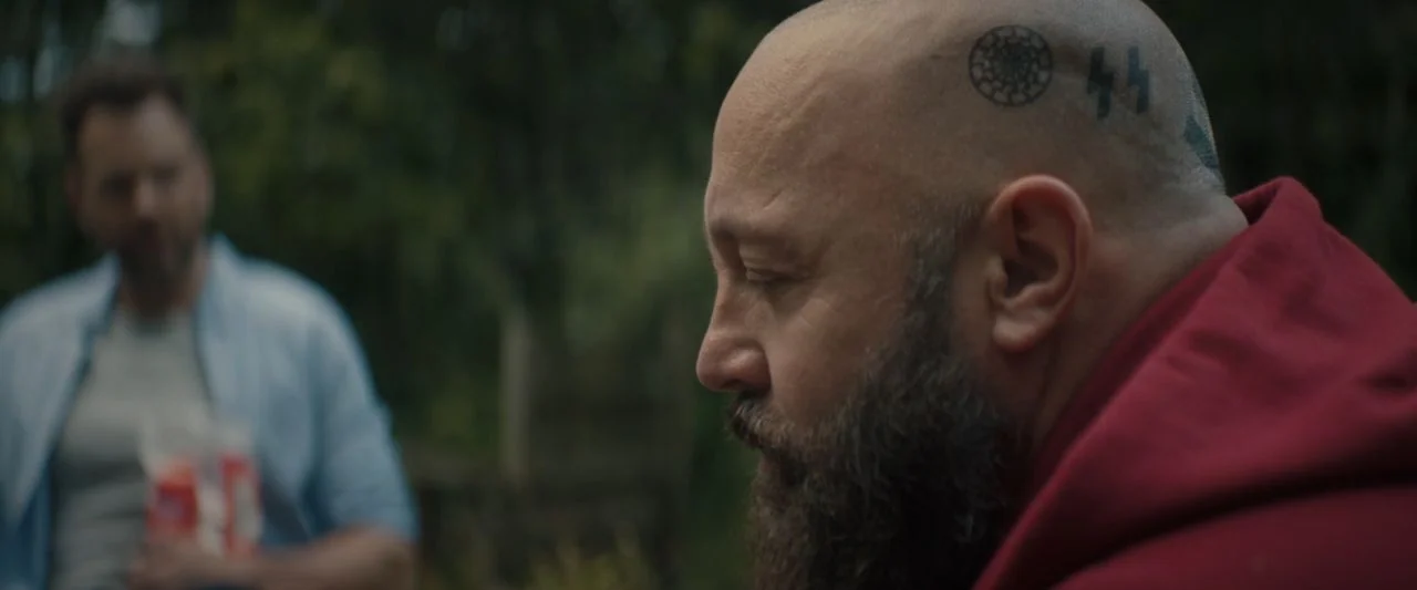 Becky (2020) movie still: medium — A bald man with a beard sits in a grassy outdoor area with trees visible. A man in a blue shirt stands in the…