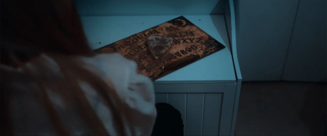 Anomaly (2006) movie still: medium — A dimly lit room features a worn wooden ouija board resting on a white table. A person with long red hair is…