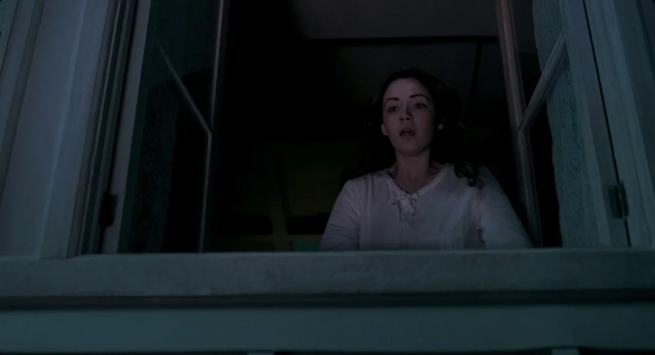 The Moth Diaries (2011) movie still: medium — A dimly lit room at night looking out through an open house window. A woman peers out from the window…