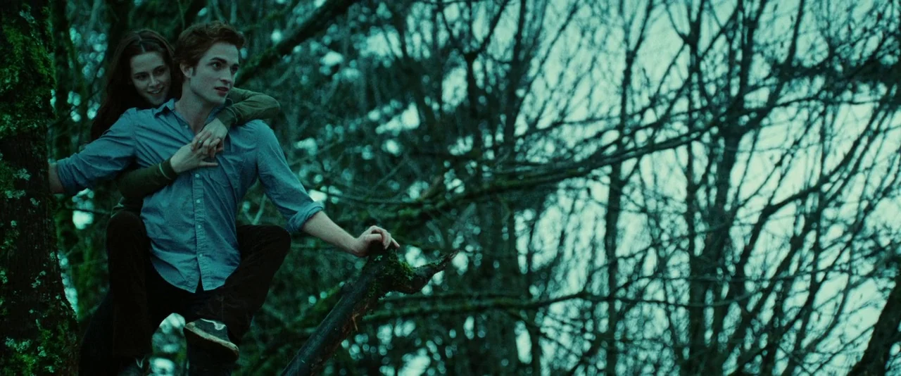 Twilight (2008) movie still: fullbody — Two individuals sit together on a thick, moss-covered tree branch high above the ground. The background is…