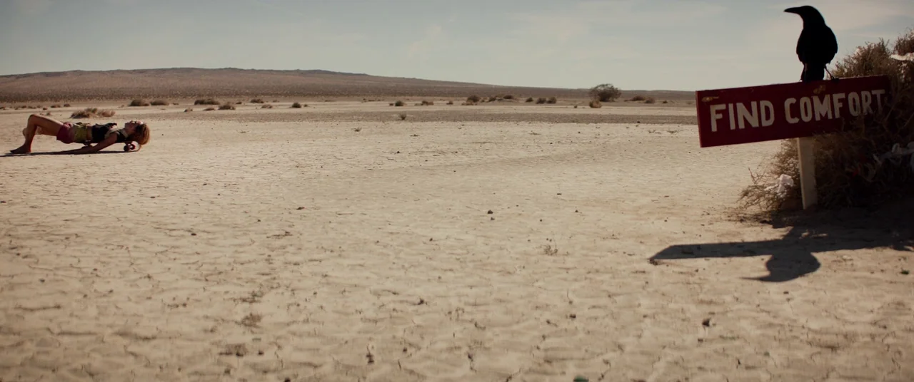 The Bad Batch (2016) movie still: wide — A vast, cracked dry lake bed stretches toward low hills under a clear sky. A lone woman lies on a…
