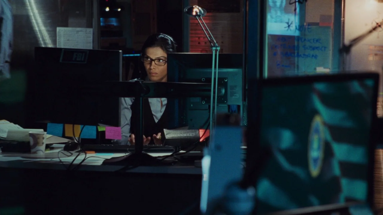 Saw V (2008) movie still: medium — A dimly lit office workspace features rows of computer monitors and handwritten notes on glass. A woman in a vest…