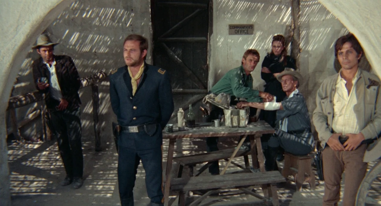 The Hellbenders (1967) movie still: fullbody — A group of six people stand within a sun-drenched, rustic military outpost courtyard. They observe a…