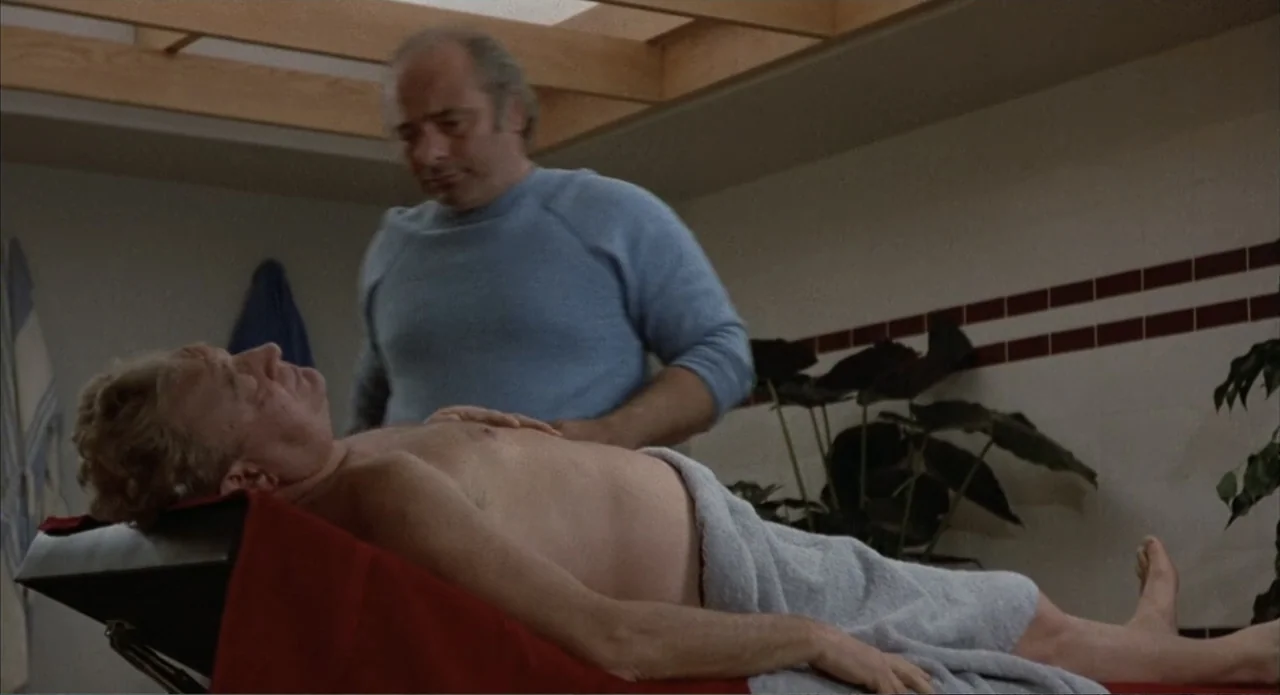 Back to School (1986) movie still: medium — An indoor room contains a massage table under an architectural slatted skylight. A man in a blue sweater…