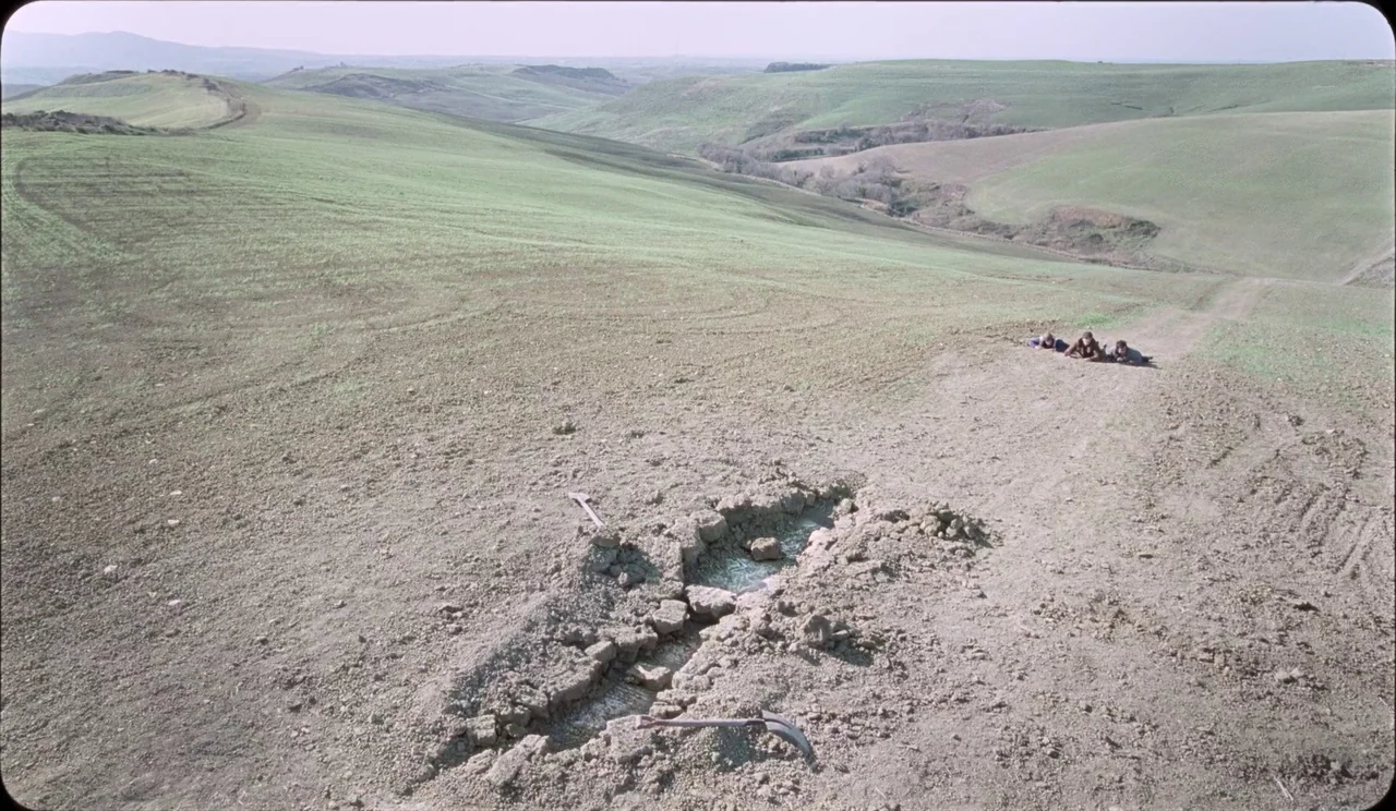 La Chimera (2023) movie still: wide — A vast, rolling green countryside with a narrow dirt path cutting through the hills. Three men lie on the…