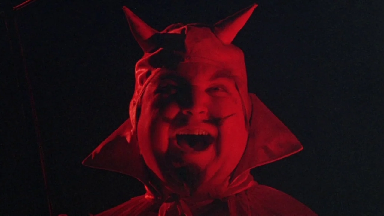 Pee-Wee’s Big Adventure (1985) movie still: closeup — A man wearing a red fabric devil costume stands against a dark background. He is laughing with…