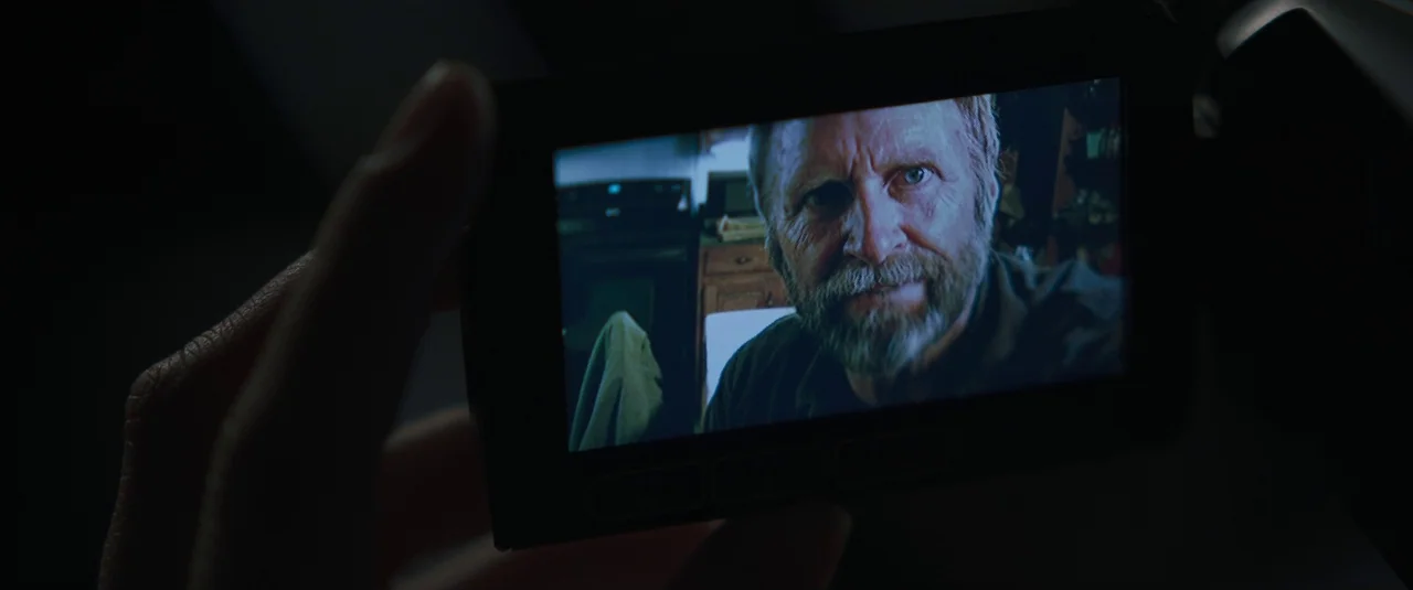 Intrusion (2021) movie still: closeup — A handheld video camera display shows a middle-aged man looking into the lens. The background within the…
