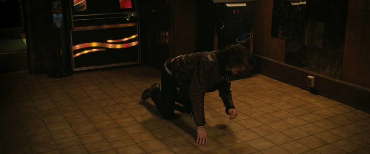 For Ellen (2012) movie still: fullbody — A dimly lit room with square brown tiles and a dark, illuminated jukebox feature. A young man kneels on the…
