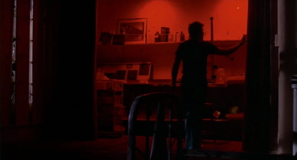 Cat People (1982) movie still: medium — A darkroom bathed in dim red light contains shelves filled with chemical bottles. A man stands silhouetted…