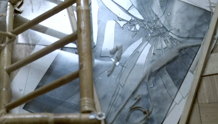 The Seventh Continent (1989) movie still: closeup — A shattered mirror lies facedown on a light-colored wooden floor with parquet patterns. A bamboo…