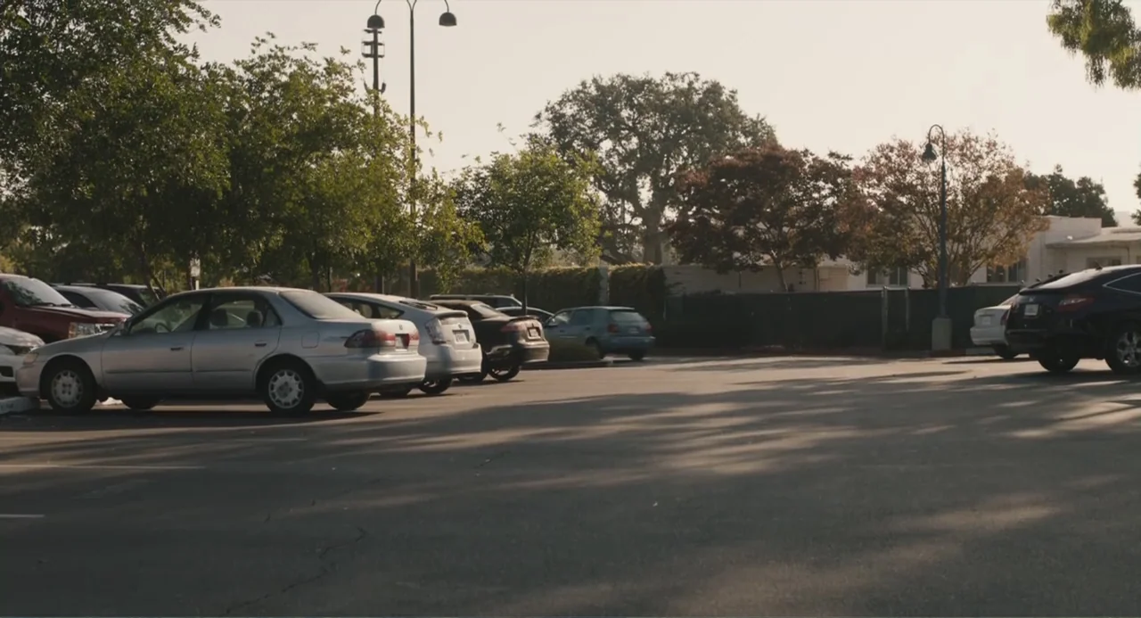 Beatriz at Dinner (2017) movie still: wide — An asphalt parking lot filled with parked sedans sits under a bright sky. Numerous leafy green trees…
