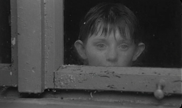 Sátántangó (1994) movie still: closeup — A dark, gritty window frame reveals a young boy looking through the glass. The child peers from the…