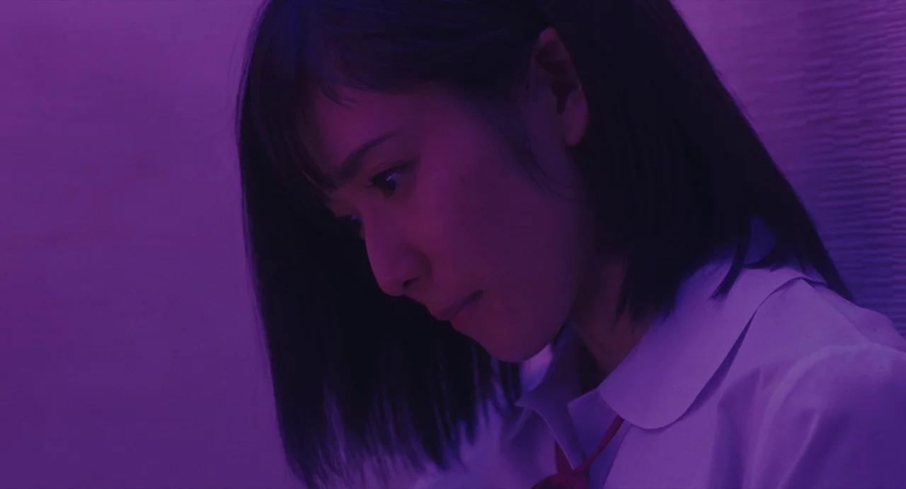 Shoplifters (2018) movie still: closeup — A dimly lit interior space is bathed in a distinct purple light. A young woman with shoulder-length black…