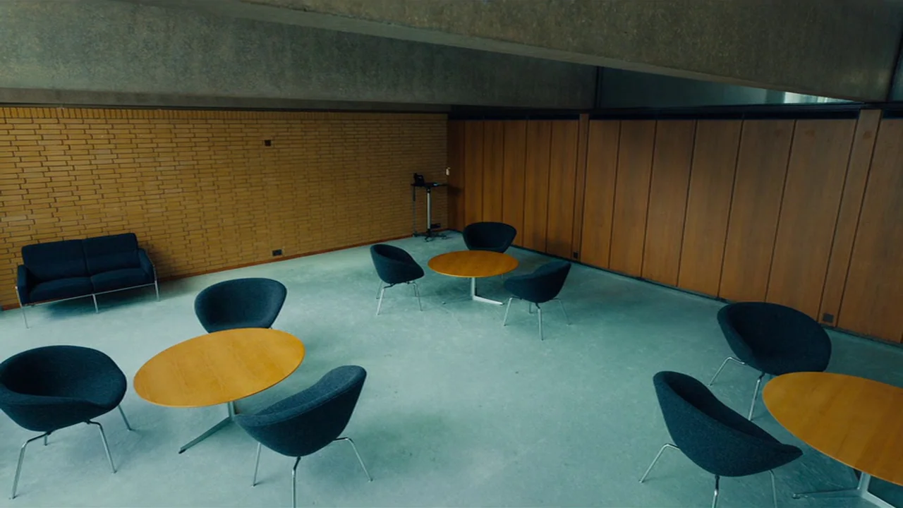 Club Zero (2023) movie still: wide — A mid-century modern lounge features a yellow brick wall and dark wood paneling. Several circular wooden tables…