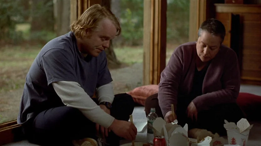 Love Liza (2002) movie still: medium — A living room interior features large floor-to-ceiling windows looking out onto a wooded area. A man and a…