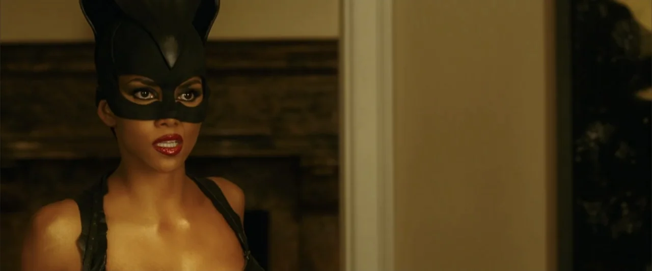 Catwoman (2004) movie still: medium — An interior living room with a stone fireplace visible in the blurred background. A woman in a black cat mask…