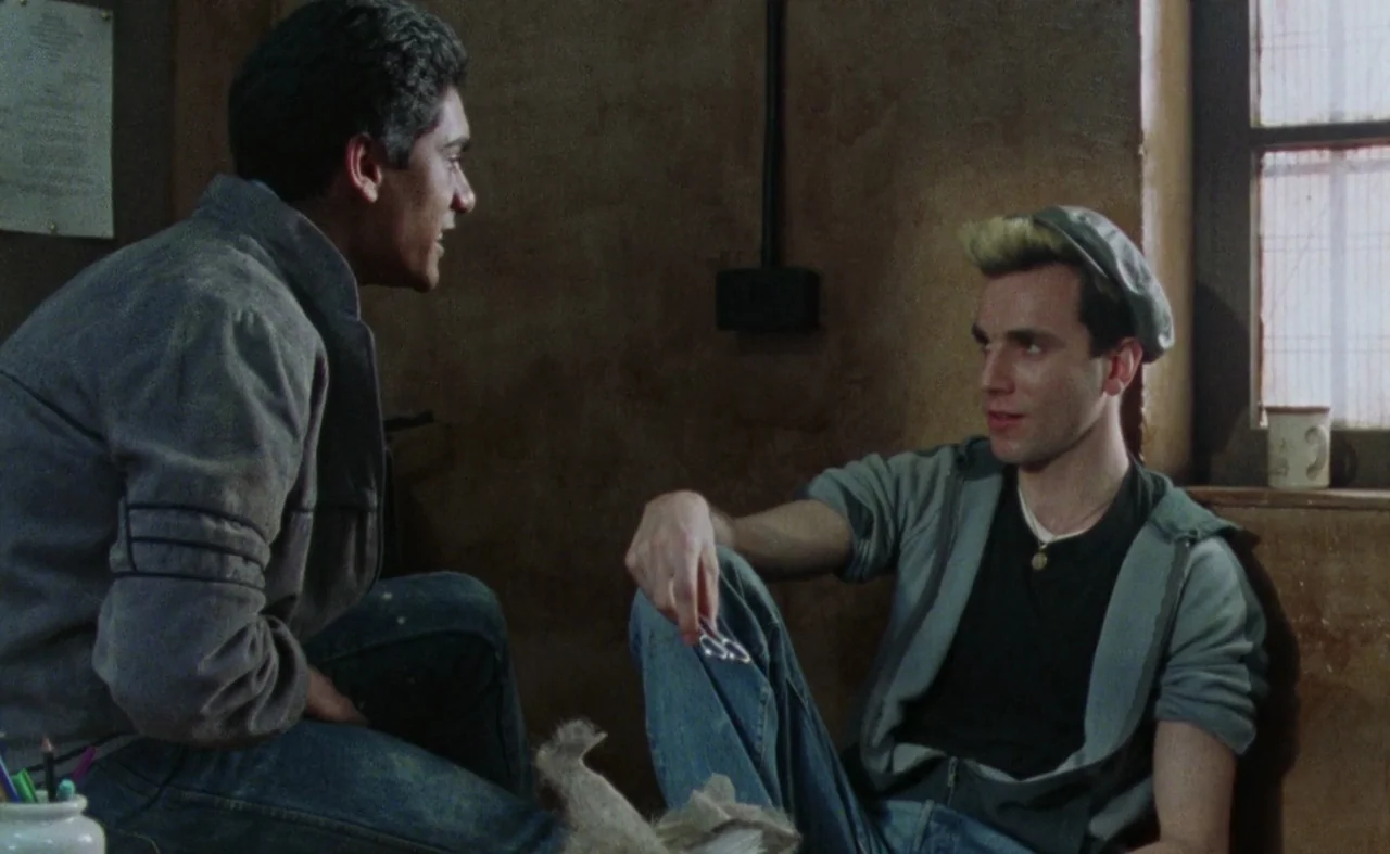 My Beautiful Laundrette (1985) movie still: medium — A dimly lit room with plain, textured walls and a simple window frame. Two young men are…