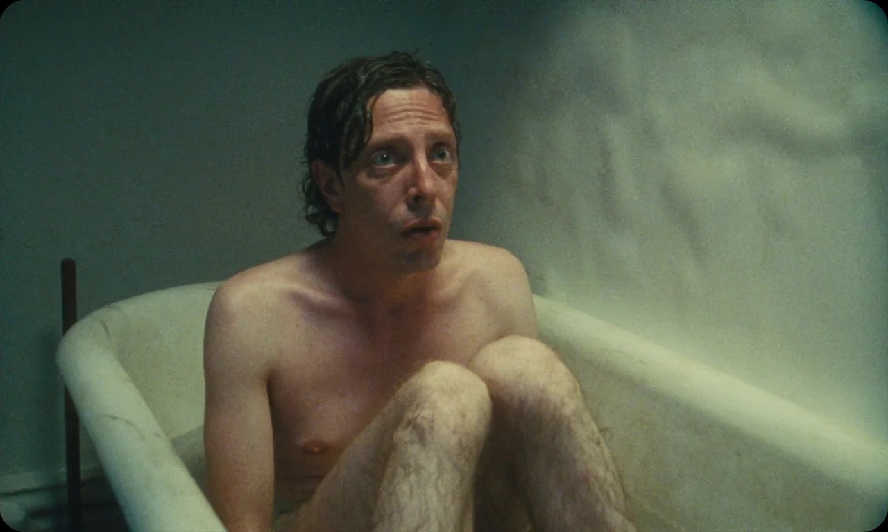 Vulcanizadora (2024) movie still: medium — A dimly lit washroom features a pale, weathered porcelain tub against a textured wall. A shirtless man…