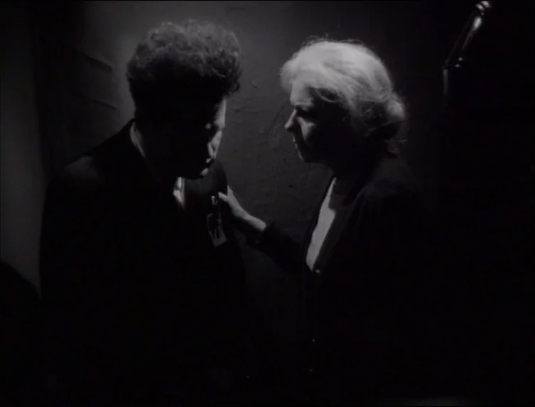 Eraserhead (1977) movie still: medium — A dimly lit, shadowed space provides a stark backdrop for a close interaction. A man stands still as an…