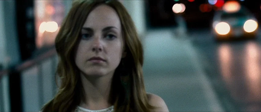 This Movie Is Broken (2010) movie still: medium — A night city street features blurred vehicular headlights in the distant background. A young woman…