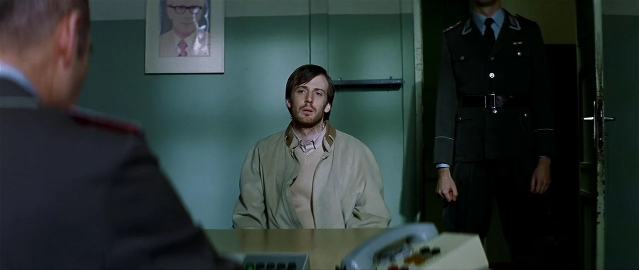 The Lives of Others (2006) movie still: medium — A spartan room features pale green walls adorned with a framed photograph of a man. A man in a…