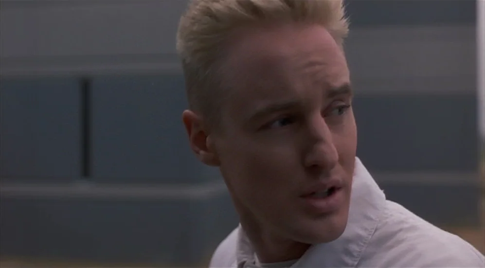 Bottle Rocket (1996) movie still: closeup — An exterior industrial location with a gray textured wall in the background. A man with short blonde…