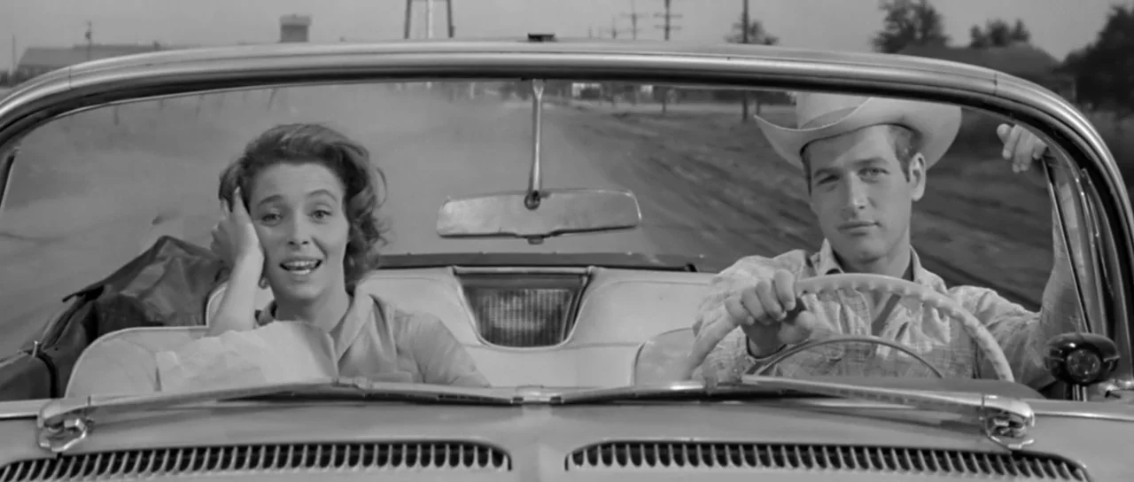 Hud (1963) movie still: medium — A man and a woman sit inside a vintage convertible driving along a road. The man drives with one hand on the…