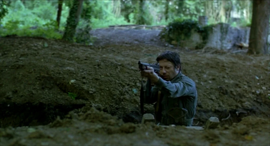 On War (2008) movie still: medium — A muddy trench surrounded by dense green woodland trees in a wartime setting. A man in a military uniform holds…